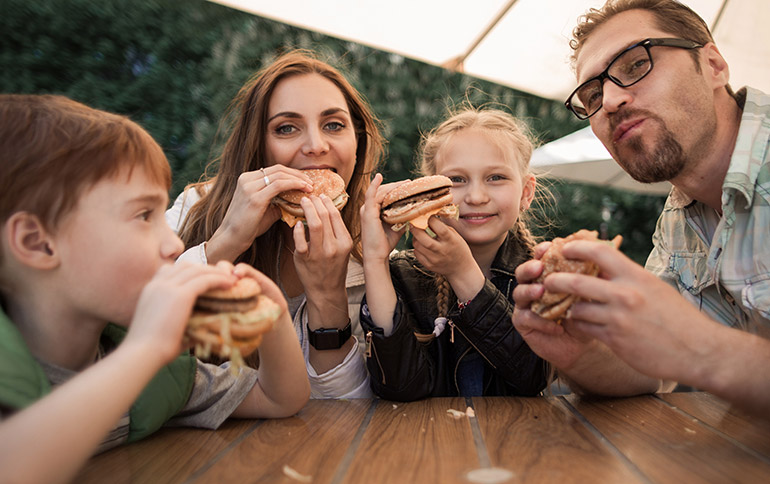 Família composta por 4 pessoas, um pai, uma mãe, dois filhos, uma menina e um menino de aproximadamente 10 anos de idade comendo hambúrgueres em uma mesa externa de madeira coberta por um guarda sol.