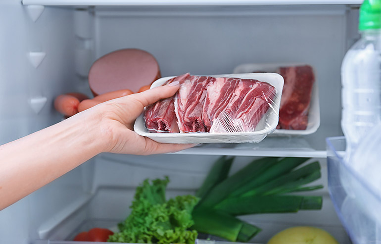Mão com uma bandeja embalada de carne crua abrindo o congelador para guardar a carne dentro